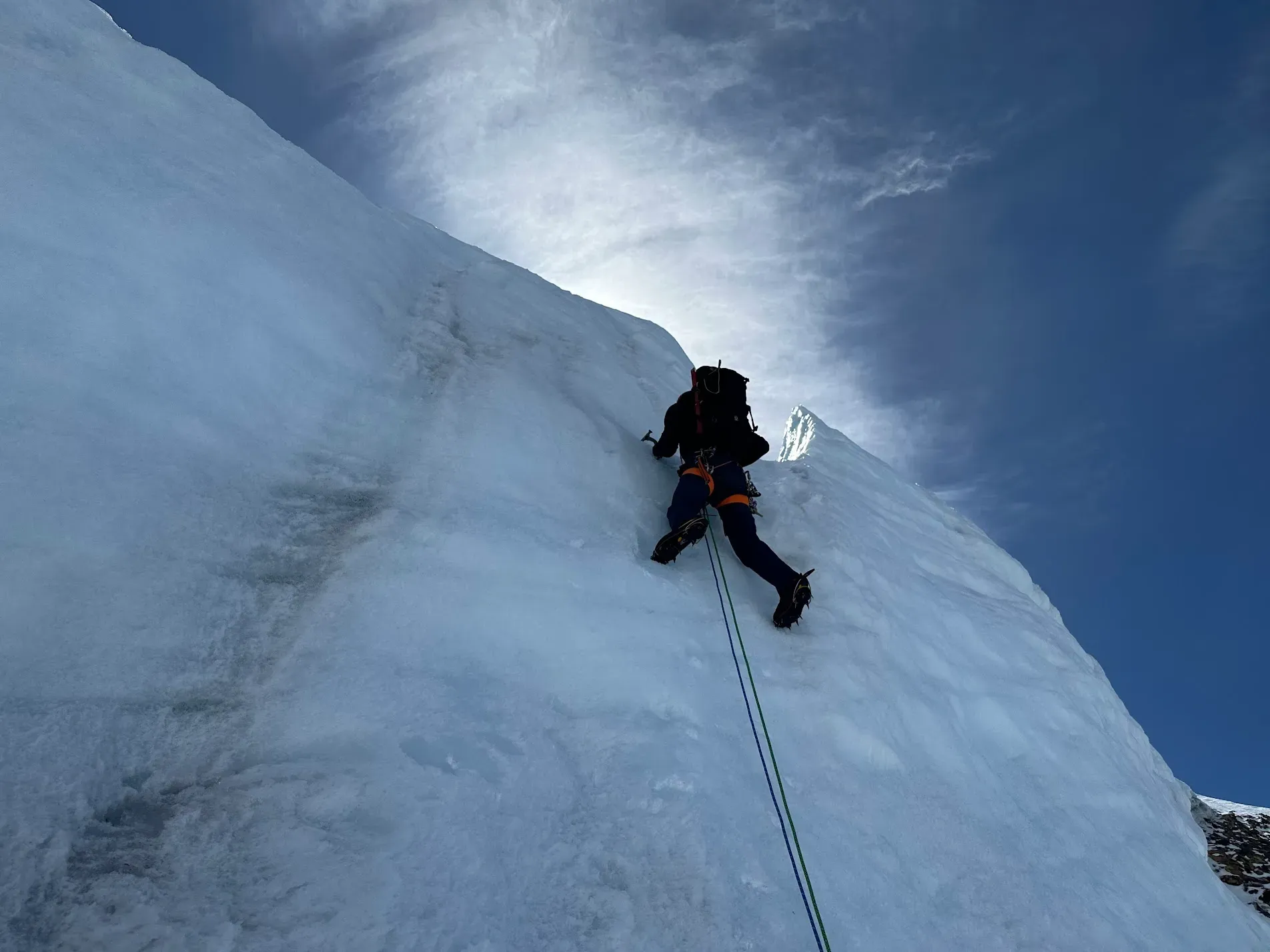 A climber ascends a section of vertical ice climbing, two ropes hang below, and a blue sky opens above