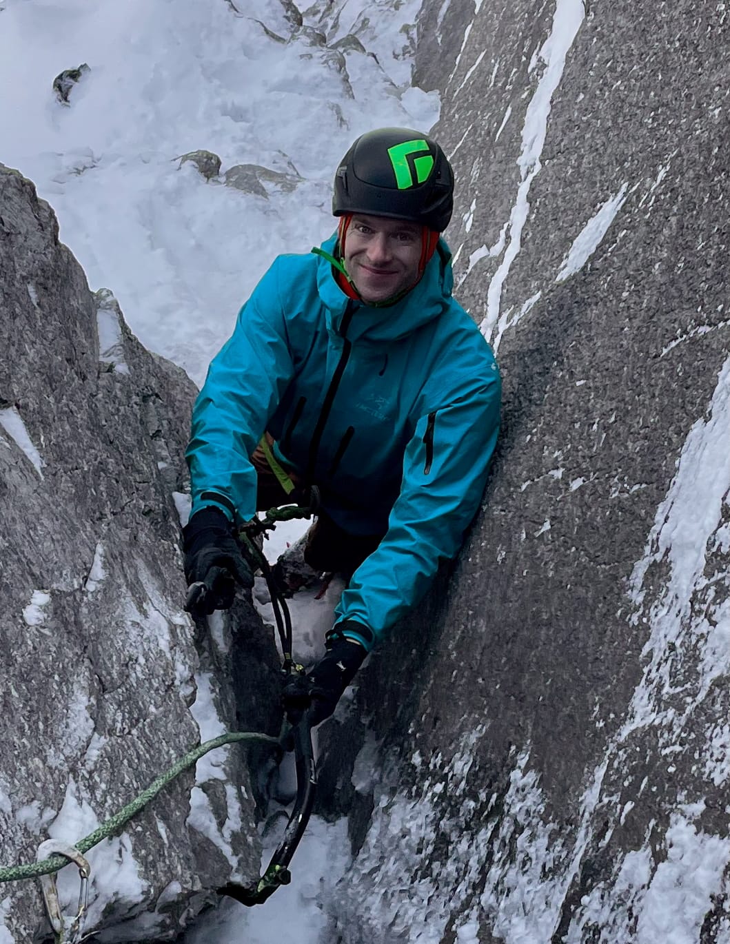 Greg Barrett, ice tools in hand, climbs a narrow corner of snowy rock. He wear a blue jacket and black helmet, and is looking at the camera, smiling.