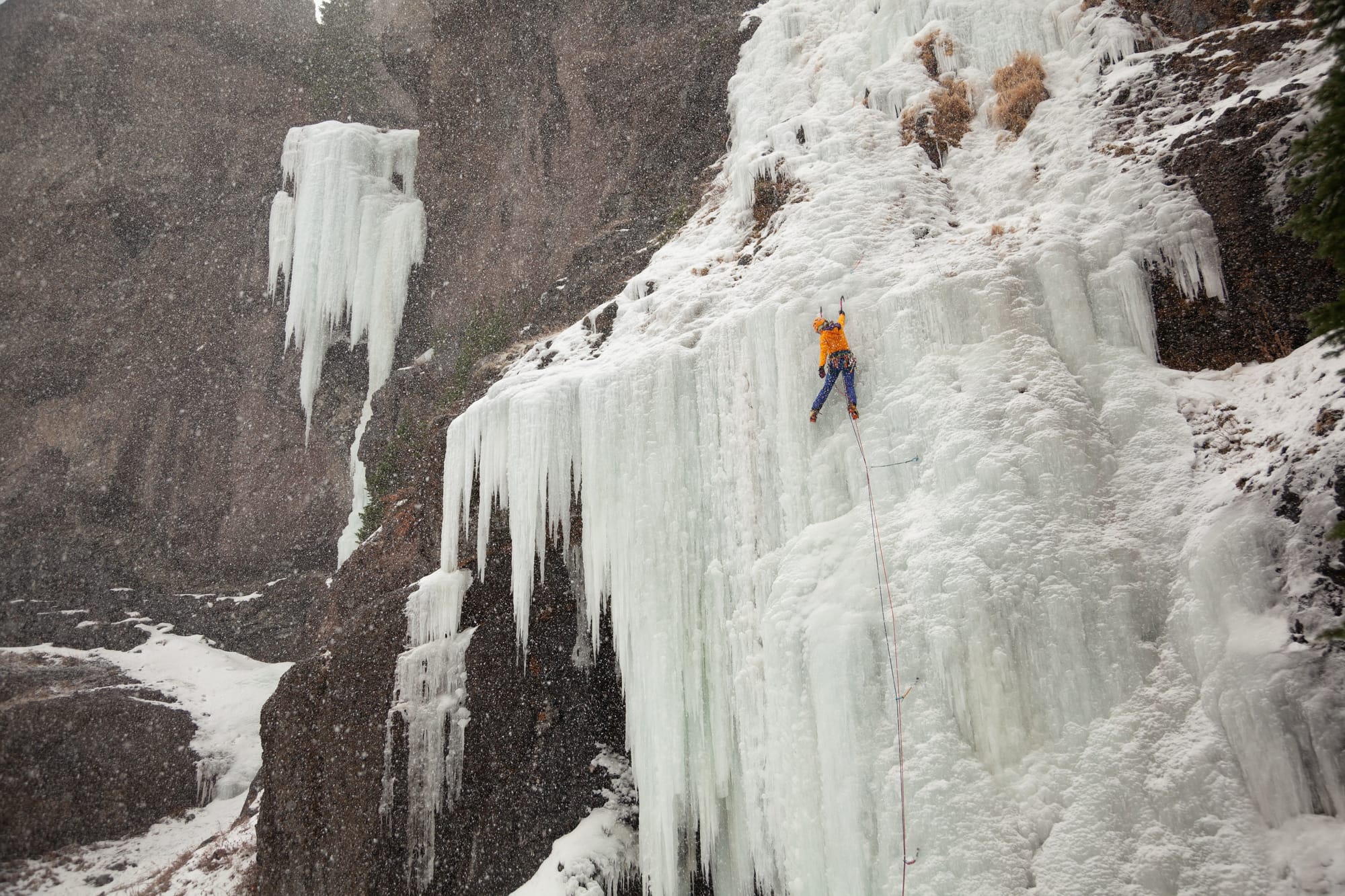 Katie McKinstry Stylos, wearing an orange jacket, leads a section of vertical ice climbing. Two ropes drape below her, connected to three ice screws.