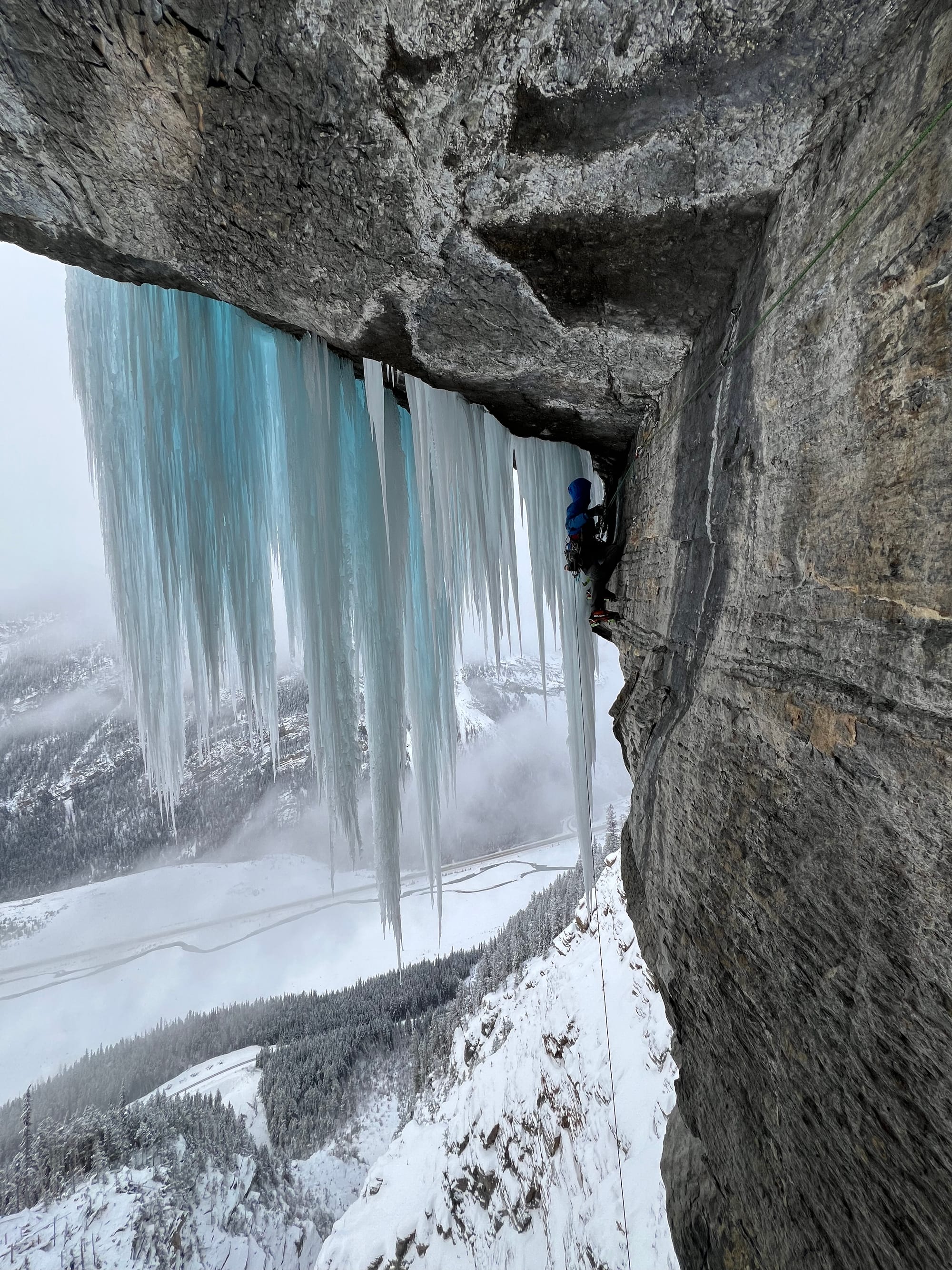 High off the ground, a winter climber makes a horizontal traverse on blank rock towards a huge sheet of hanging icicles. 