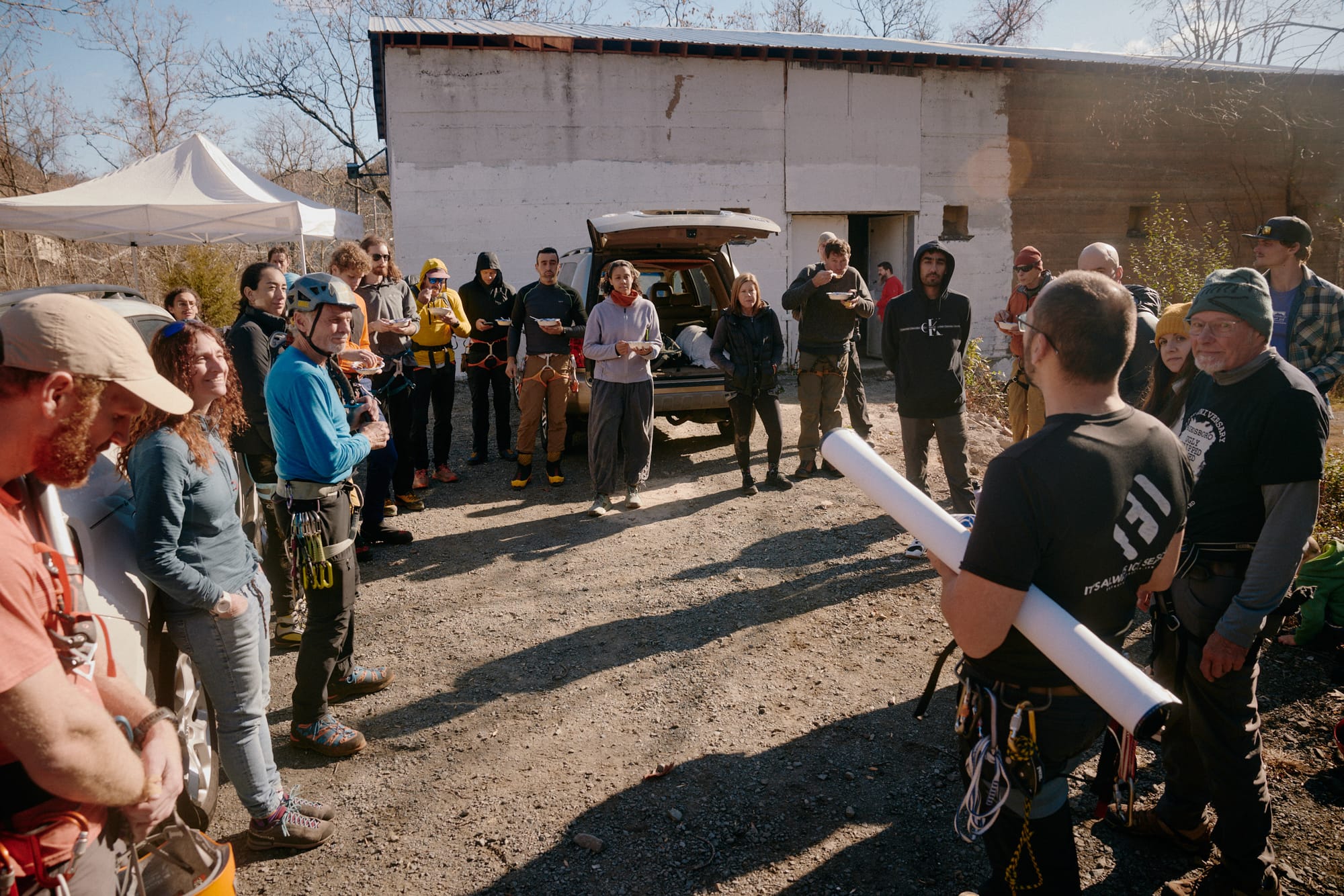 The 2024 Birdsboro Drytool Enclave gathers for lunch; more than people stand in a loose circle, some hold bowls from which they are eating while others look at one man who is speaking