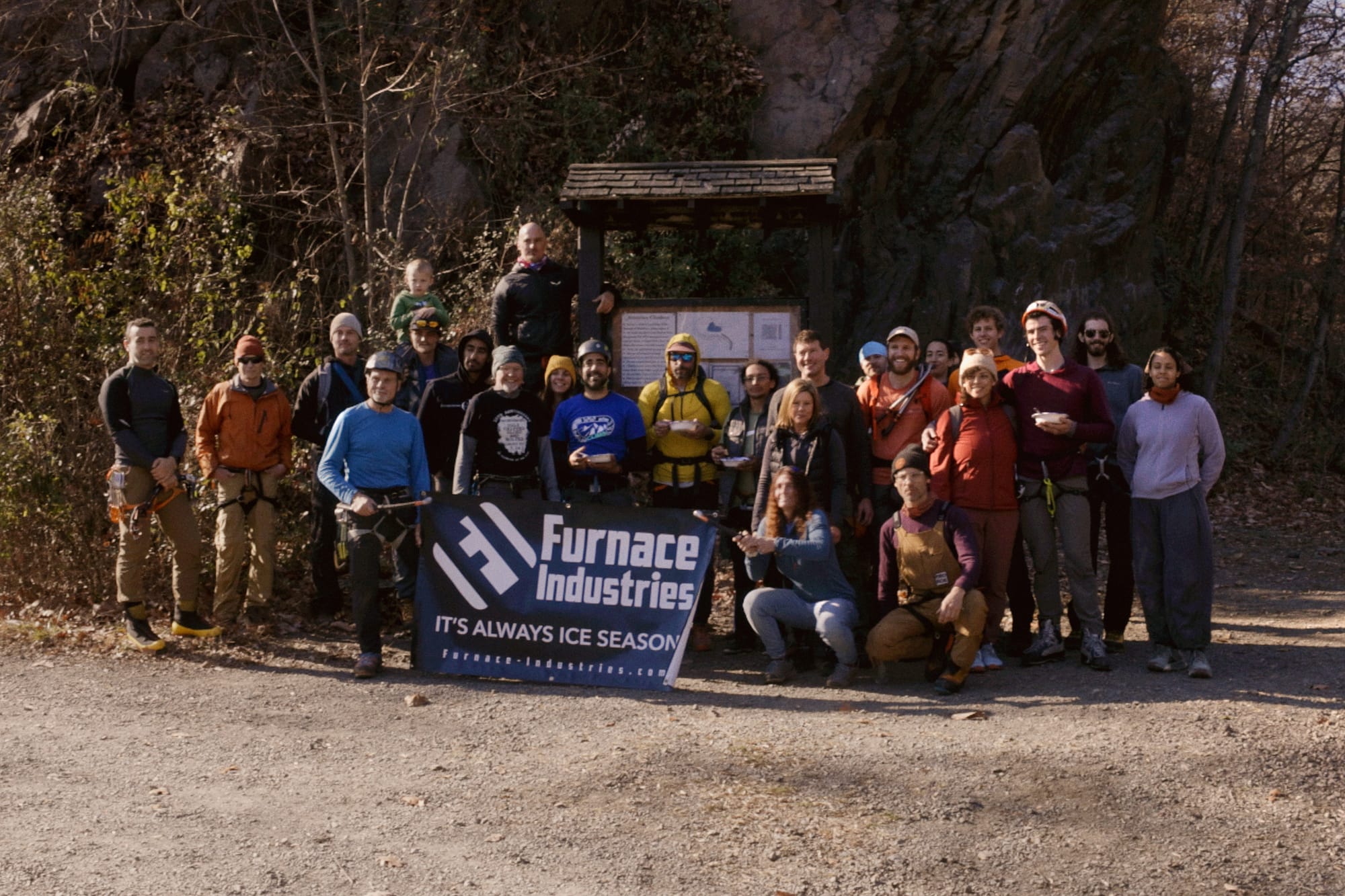 Group photo of participamnts at the Fall 2024 Birdsboro Drytool Enclave, posing in front of a trailhead sign with a banner from the event's main sponsor, Furnace Industries